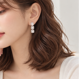 A woman with shoulder-length brown wavy hair wears a delicate silver drop earring with three clear gemstones, posing in a bright, softly lit indoor setting.