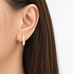 A close-up of a model's ear wearing a small gold huggie hoop earring with clear baguette-cut stones, set against a clean white background.