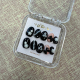 Set of black and white nail art samples in a clear display case on a textured surface.