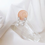 A pair of silver drop earrings with clear gemstones, resting on a clear glass block next to a peach wax seal stamp, set against a soft white draped fabric background.