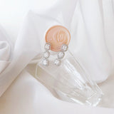 A pair of silver drop earrings with clear gemstones, resting on a clear glass block next to a peach wax seal stamp, set against a soft white draped fabric background.