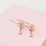 A pair of rose gold butterfly dangle earrings with clear rhinestones, resting on a textured light pink gift box with gold foil script.