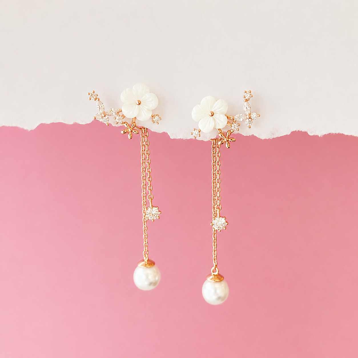 A pair of gold-tone dangle earrings with white flower charms, small clear crystal accents, and white pearl drops, displayed against a pink and white split background.