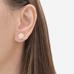 A close-up of a model's ear wearing a delicate floral ear cuff earring in rose gold with white flower petals and small clear stones, set against a clean white background.