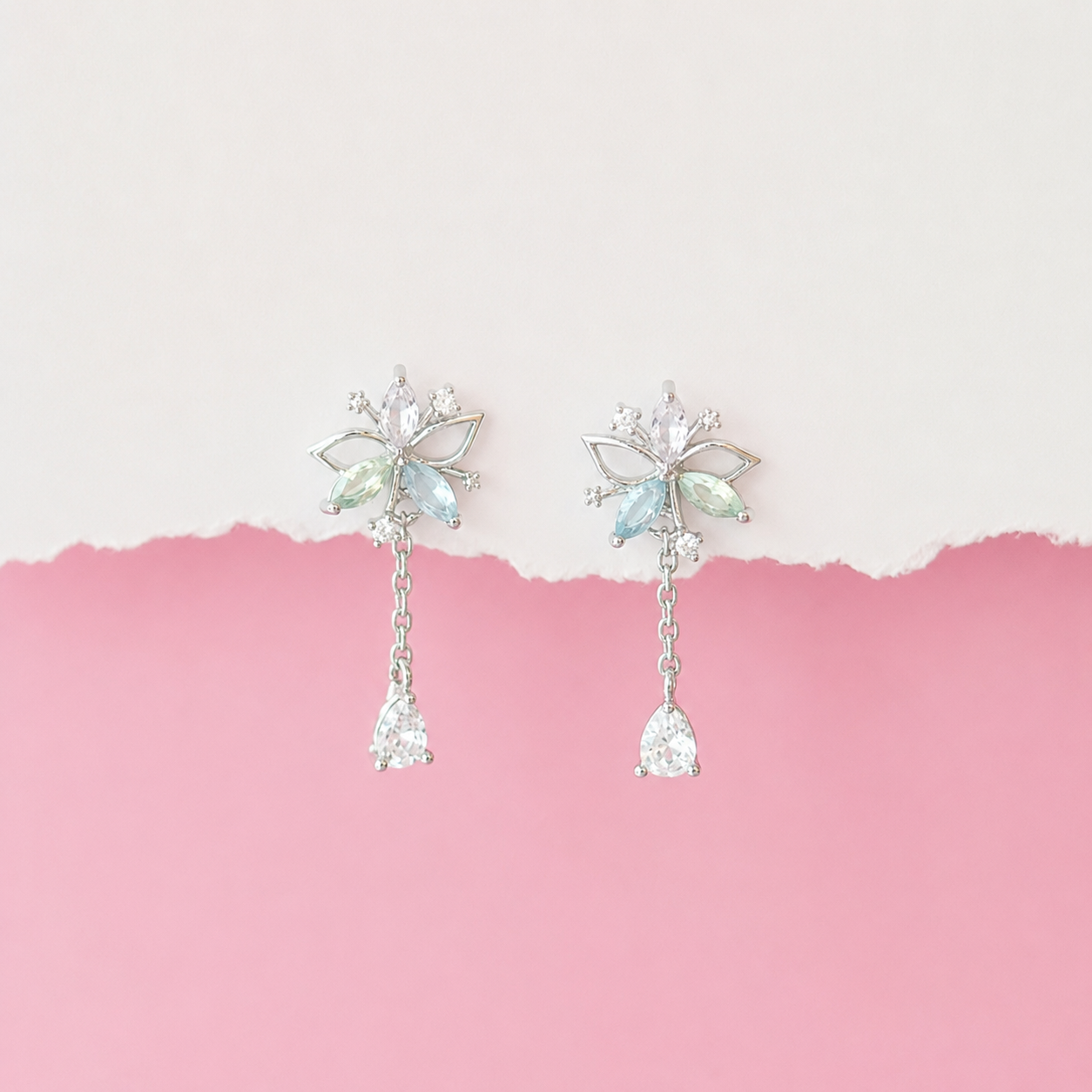 A pair of silver-toned dangle earrings with iridescent light blue and green crystal details, displayed against a split white and pink background.