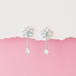 A pair of silver-toned dangle earrings with iridescent light blue and green crystal details, displayed against a split white and pink background.