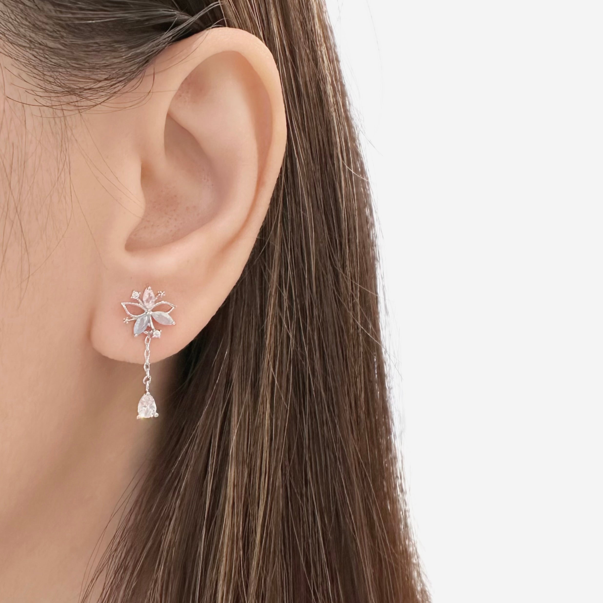A close-up of a model's ear wearing a silver-toned dangle earring with a butterfly-shaped top and a small clear crystal bell hanging below.