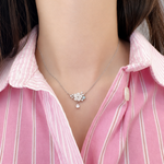 A close-up of a model wearing a delicate silver necklace with a small floral cluster and teardrop pendant, styled with a pink and white striped button-down shirt.