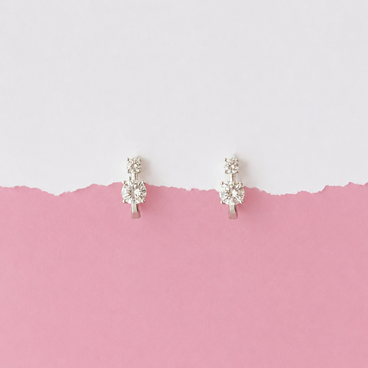 A pair of small silver-toned hoop earrings with clear gemstones, displayed against a split white and pink background.