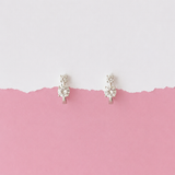A pair of small silver-toned hoop earrings with clear gemstones, displayed against a split white and pink background.