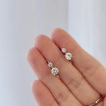 A close-up of a hand holding a pair of small silver stud earrings with clear round-cut gemstones, against a soft light gray background.