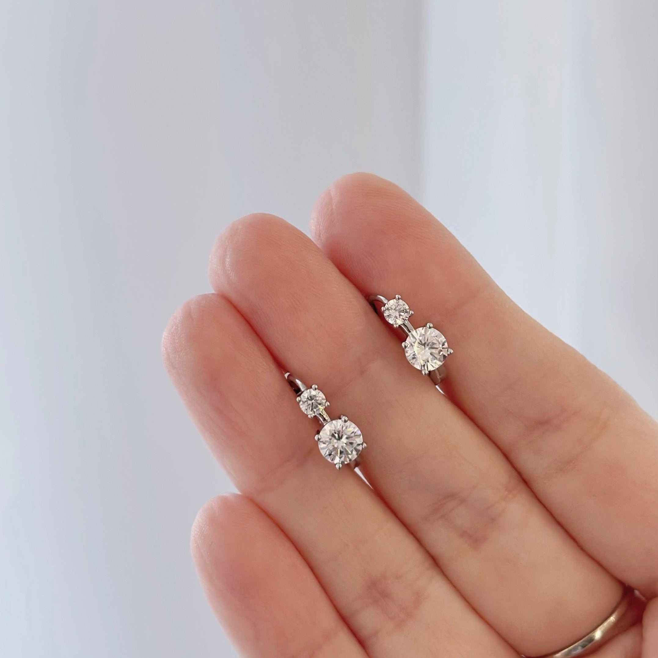 A close-up of a hand holding a pair of small silver stud earrings with clear round-cut gemstones, against a soft light gray background.