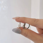 A close-up of a hand with light gray manicured nails holding two silver-toned rings with clear gemstones against a soft white background.