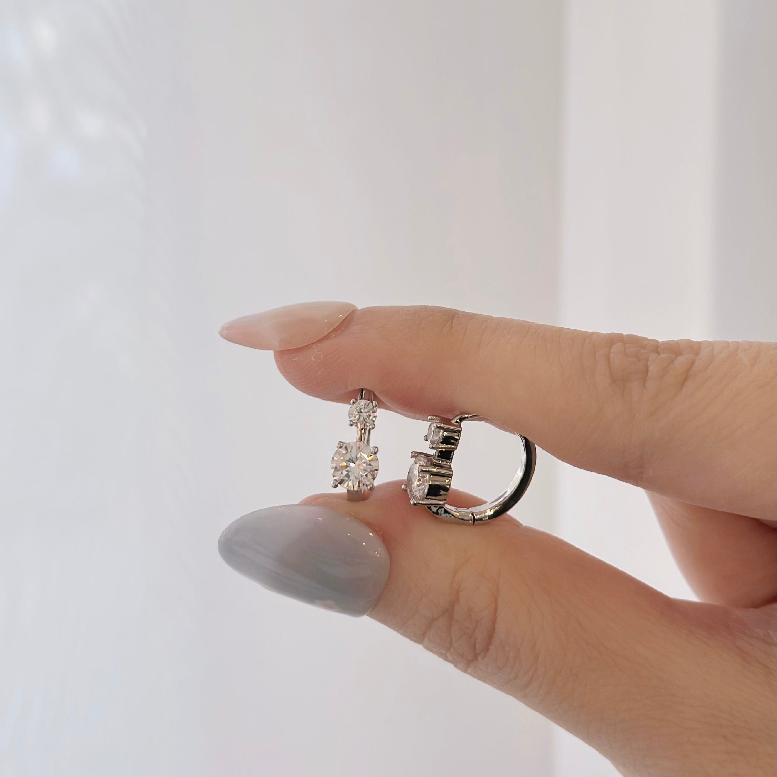 A close-up of a hand with light gray manicured nails holding two silver-toned rings with clear gemstones against a soft white background.